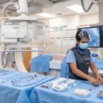 A female cath lab tech wearing a lead apron, face mask, and shield carefully organizing sterile catheters and guide wires on a blue draped table inside a cardiac catheterization laboratory.