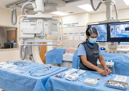 A female cath lab tech wearing a lead apron, face mask, and shield carefully organizing sterile catheters and guide wires on a blue draped table inside a cardiac catheterization laboratory.